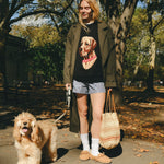 Woman wearing a black knit sweater with an intarsia dog portrait on the front and a red collar detail, walking a dog through the park. 