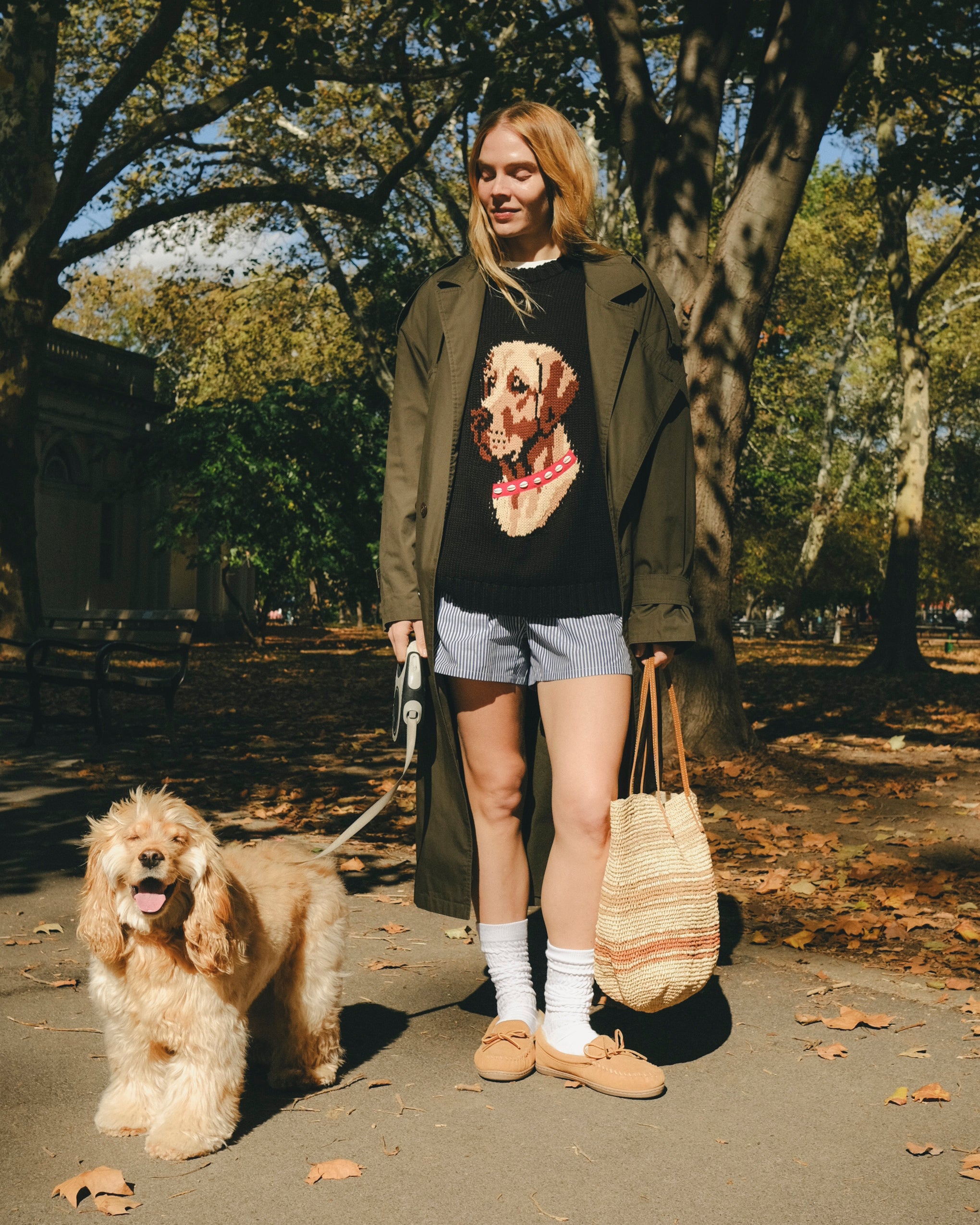 Woman wearing a black knit sweater with an intarsia dog portrait on the front and a red collar detail, walking a dog through the park. 