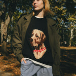 Woman wearing a black knit sweater with ribbed collar, cuffs, and hem, featuring an intarsia dog portrait on the front wearing a red collar with white shell details.