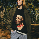 Woman wearing a black knit sweater with ribbed collar, cuffs, and hem, featuring an intarsia dog portrait on the front wearing a red collar with white shell details.