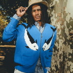 Model wearing a blue zip-up knit sweater with a fish design, standing outdoors beside a tree and holding the brim of a hat.