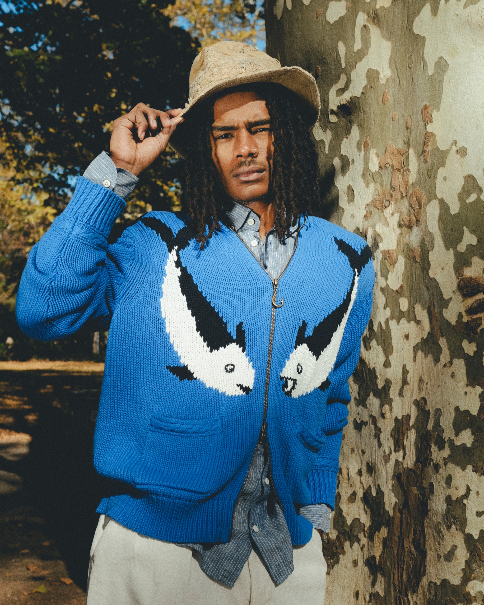 Model wearing a blue zip-up knit sweater with a fish design, standing outdoors beside a tree and holding the brim of a hat.