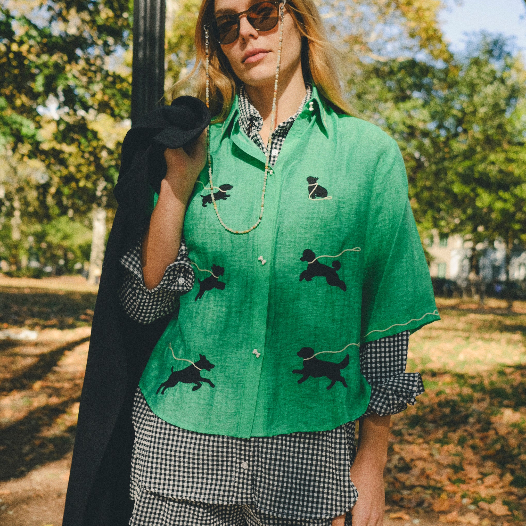 Woman wearing a green cropped cabana shirt with black dog silhouettes and white leash embroidery across the front, layered over a black-and-white gingham shirt.