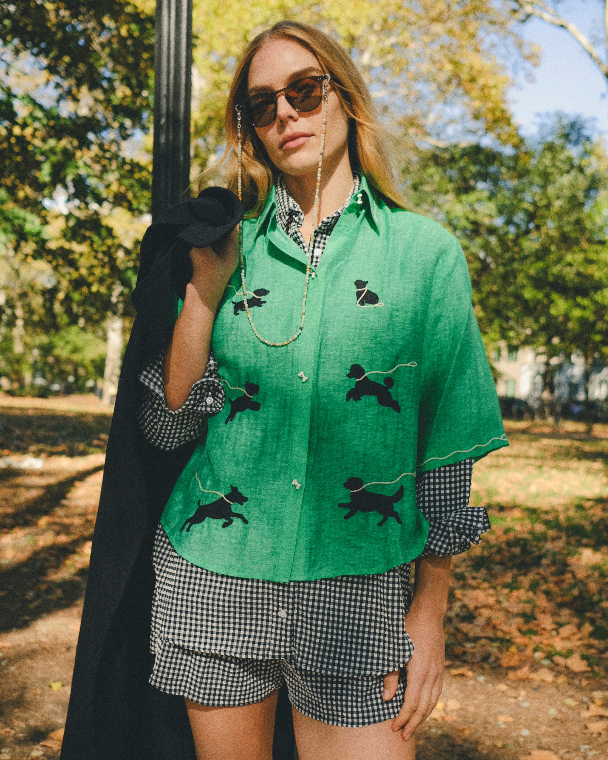 Woman wearing a green cropped cabana shirt with black dog silhouettes and white leash embroidery across the front, layered over a black-and-white gingham shirt.