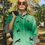 Woman wearing a green cropped cabana shirt with black dog silhouettes and white leash embroidery, layered over a black-and-white gingham shirt.