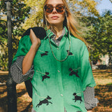 Woman wearing a green cropped cabana shirt with black dog silhouettes and white leash embroidery, layered over a black-and-white gingham shirt.