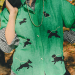 Woman wearing a green cropped cabana shirt with black dog silhouettes and white leash embroidery, layered over a black-and-white gingham shirt.