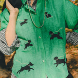 Woman wearing a green cropped cabana shirt with black dog silhouettes and white leash embroidery, layered over a black-and-white gingham shirt.