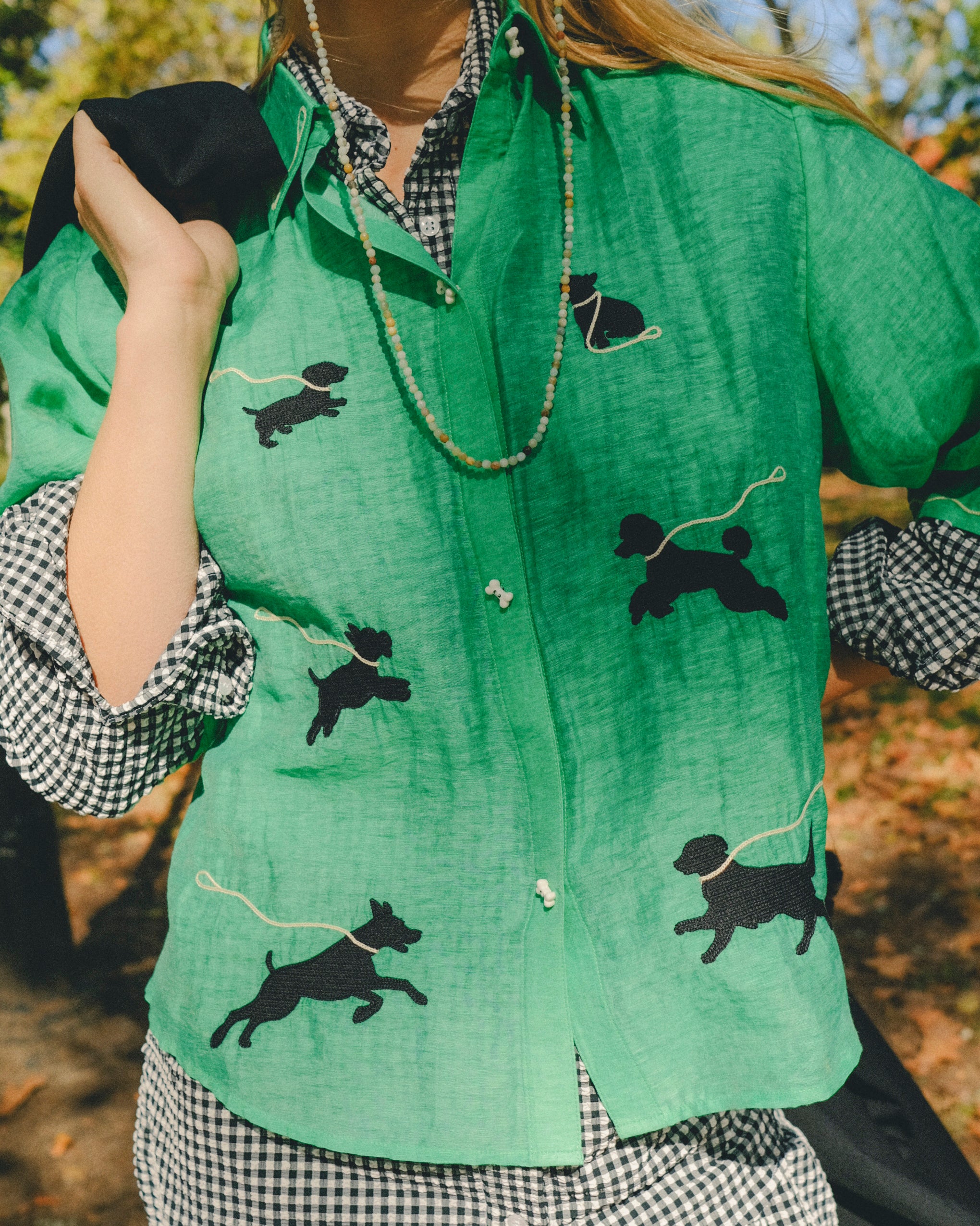Woman wearing a green cropped cabana shirt with black dog silhouettes and white leash embroidery, layered over a black-and-white gingham shirt.