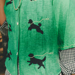 Close-up of a green cropped cabana shirt featuring black dog silhouettes connected by white leash embroidery, layered over a black-and-white gingham shirt.
