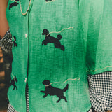 Close-up of a green cropped cabana shirt featuring black dog silhouettes connected by white leash embroidery, layered over a black-and-white gingham shirt.