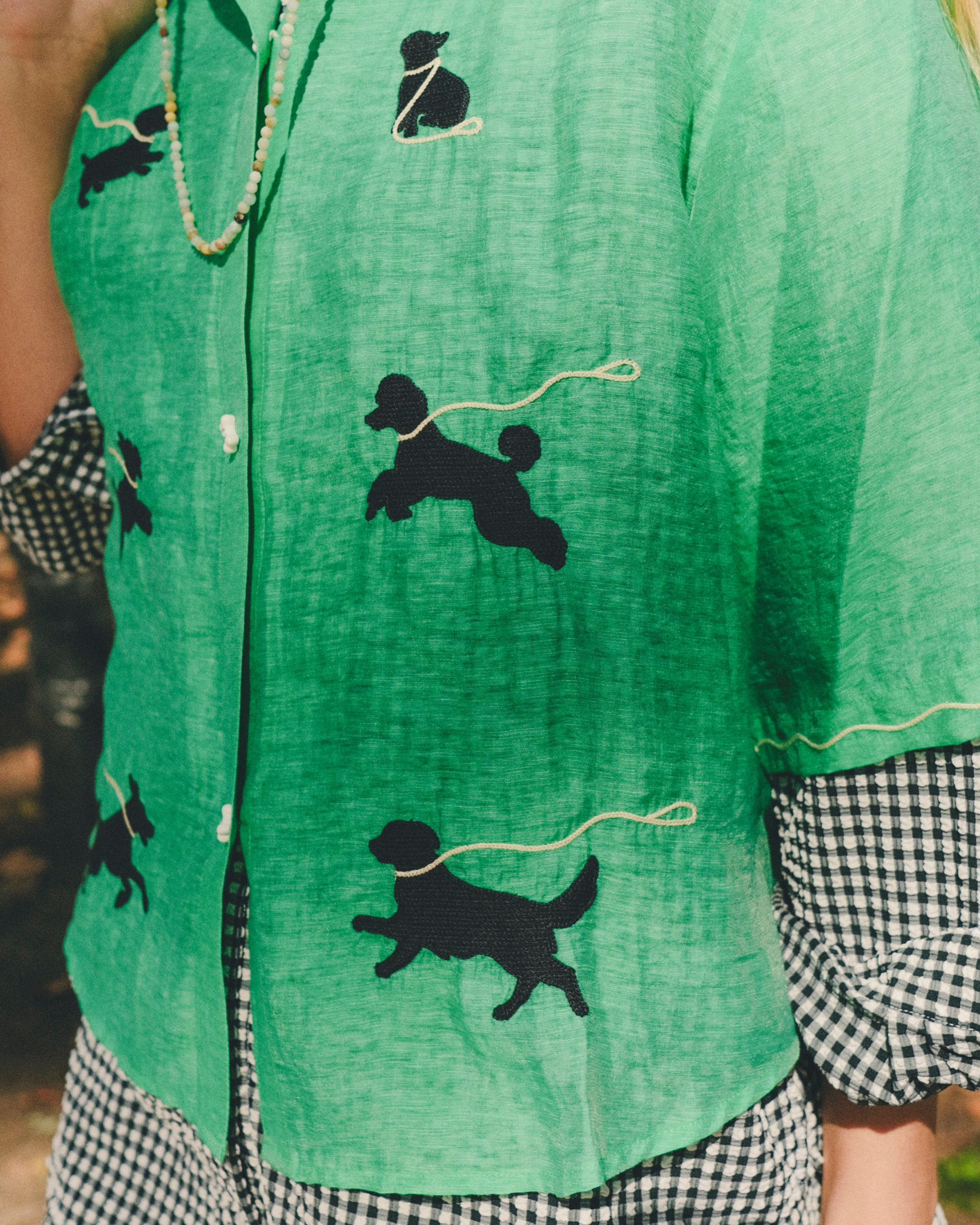 Close-up of a green cropped cabana shirt featuring black dog silhouettes connected by white leash embroidery, layered over a black-and-white gingham shirt.