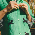 Close-up of a green cropped cabana shirt with black dog silhouettes, white leash embroidery, and dog bone–shaped buttons, layered over a black-and-white gingham shirt.
