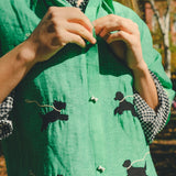 Close-up of a green cropped cabana shirt with black dog silhouettes, white leash embroidery, and dog bone–shaped buttons, layered over a black-and-white gingham shirt.