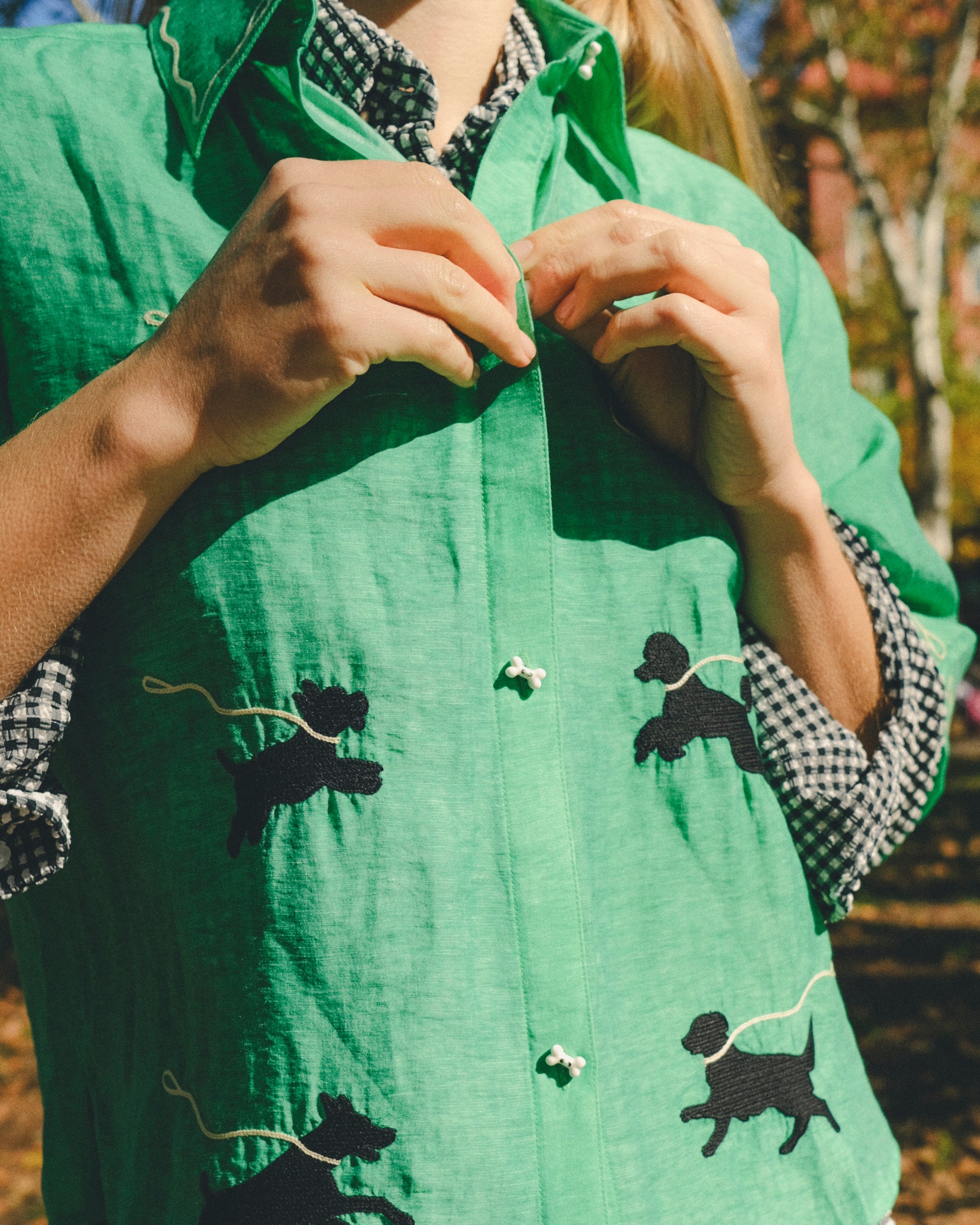 Close-up of a green cropped cabana shirt with black dog silhouettes, white leash embroidery, and dog bone–shaped buttons, layered over a black-and-white gingham shirt.