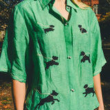 Close-up of a green cropped cabana shirt with black dog silhouettes, white leash embroidery, and dog bone–shaped buttons, layered over a black-and-white gingham shirt.