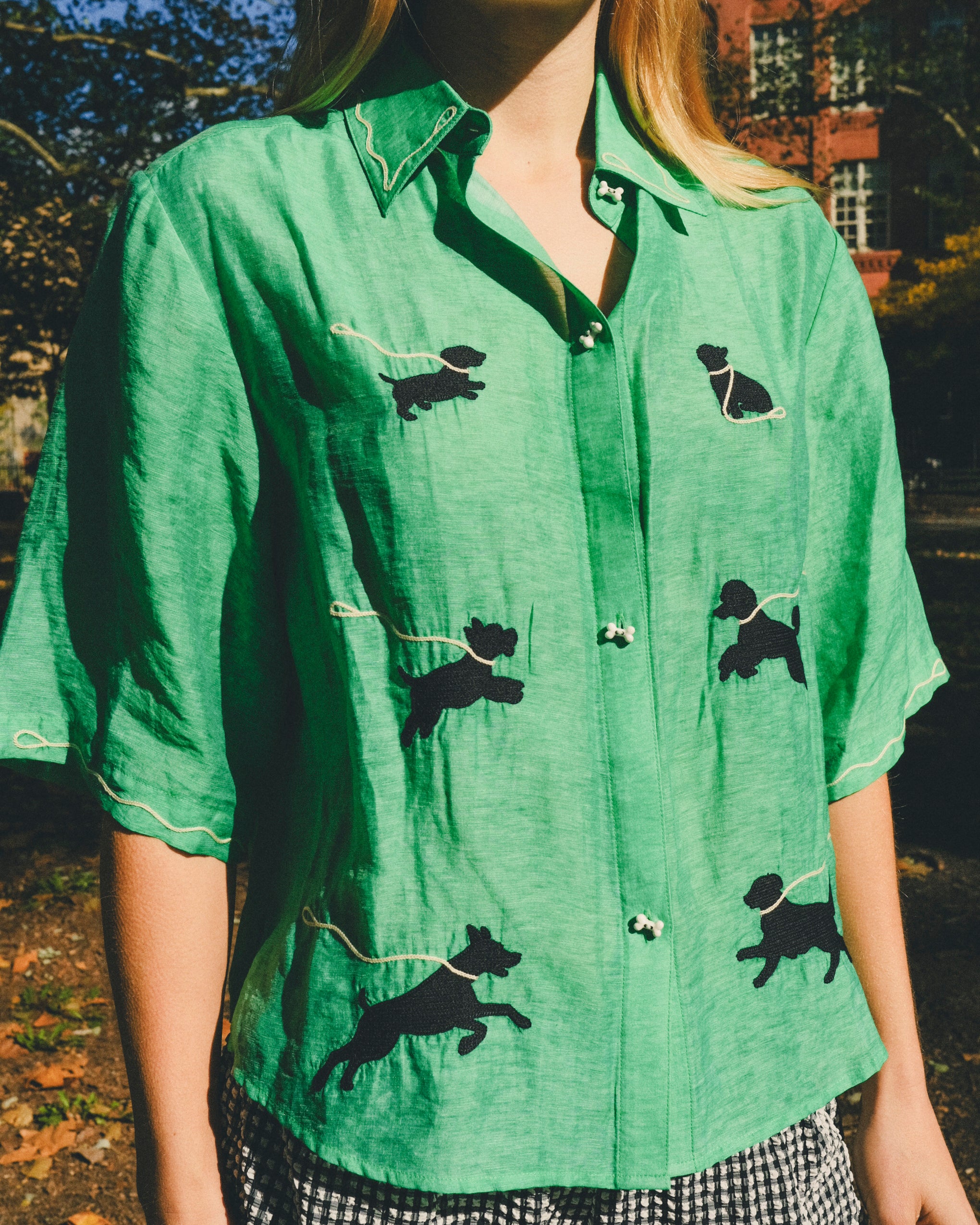 Close-up of a green cropped cabana shirt with black dog silhouettes, white leash embroidery, and dog bone–shaped buttons, layered over a black-and-white gingham shirt.