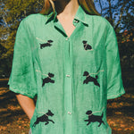 Close-up of a green cropped cabana shirt with black dog silhouettes and white leash embroidery, finished with dog bone–shaped buttons.