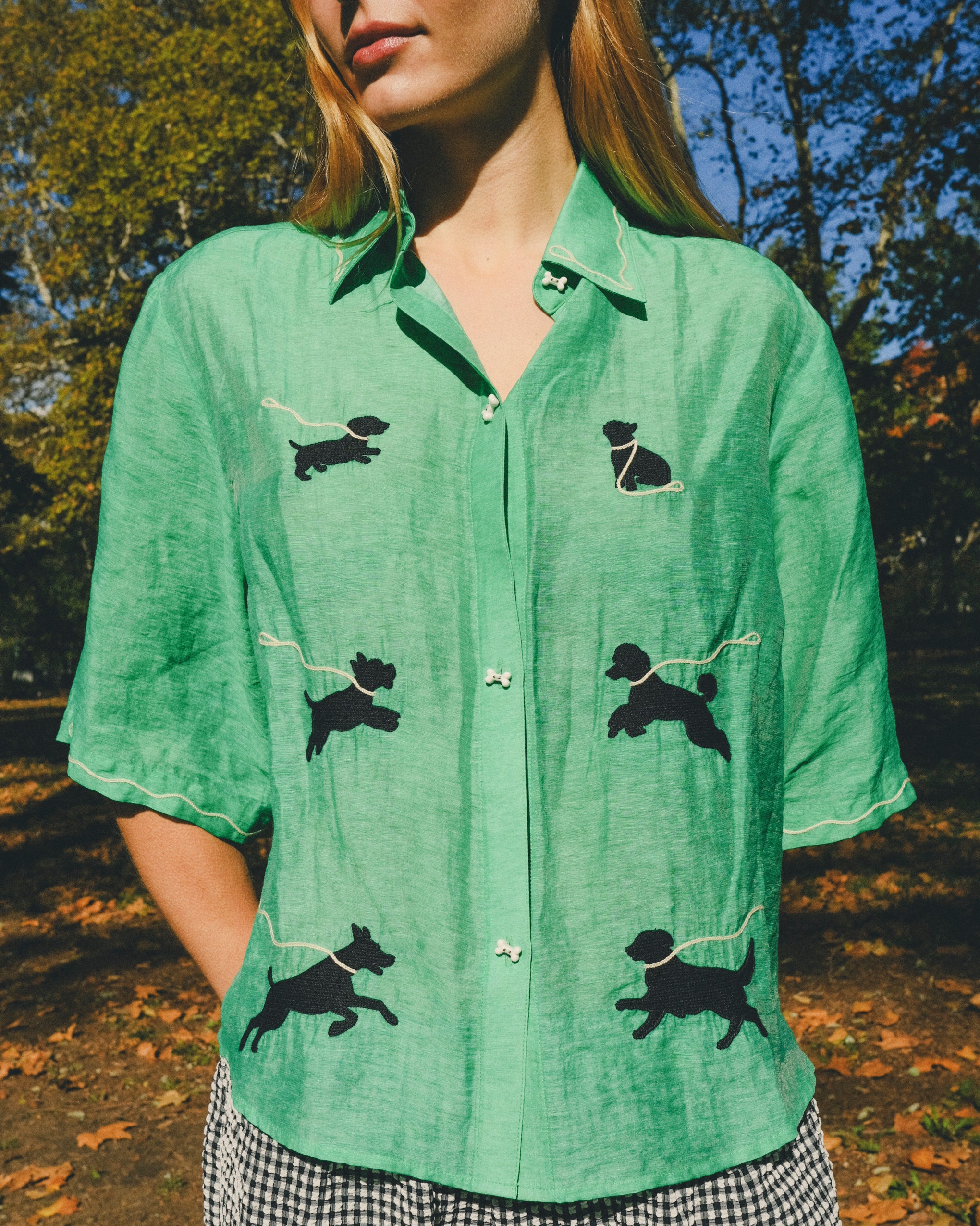 Close-up of a green cropped cabana shirt with black dog silhouettes and white leash embroidery, finished with dog bone–shaped buttons.