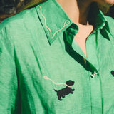 Close-up of a green cropped cabana shirt with black dog silhouettes, white leash embroidery, and dog bone–shaped buttons, layered over a black-and-white gingham shirt.