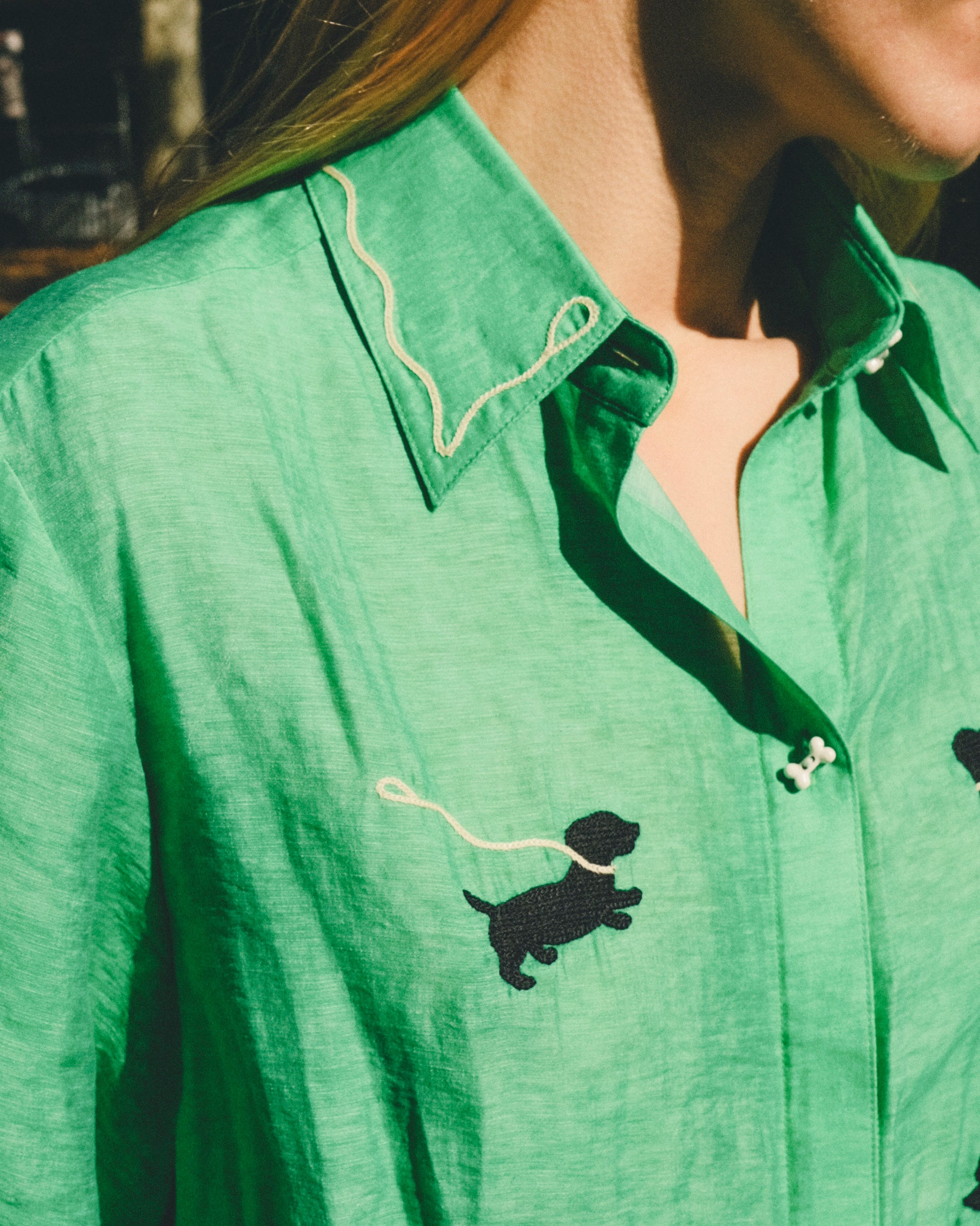 Close-up of a green cropped cabana shirt with black dog silhouettes, white leash embroidery, and dog bone–shaped buttons, layered over a black-and-white gingham shirt.