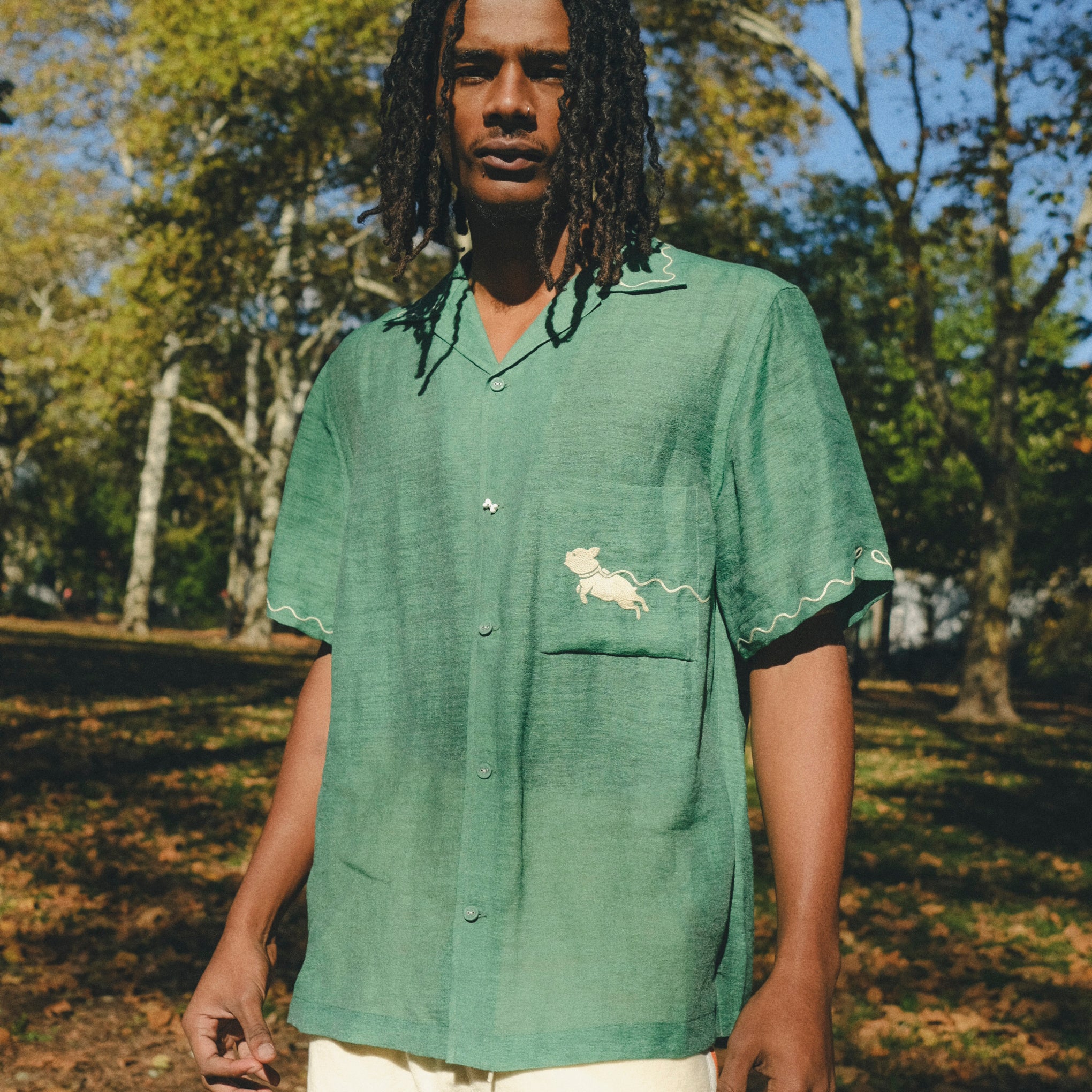 Man wearing green cabana shirt with contrast wavy piping, chest pocket with embroidered dog graphic, and a dog bone button at the center front.