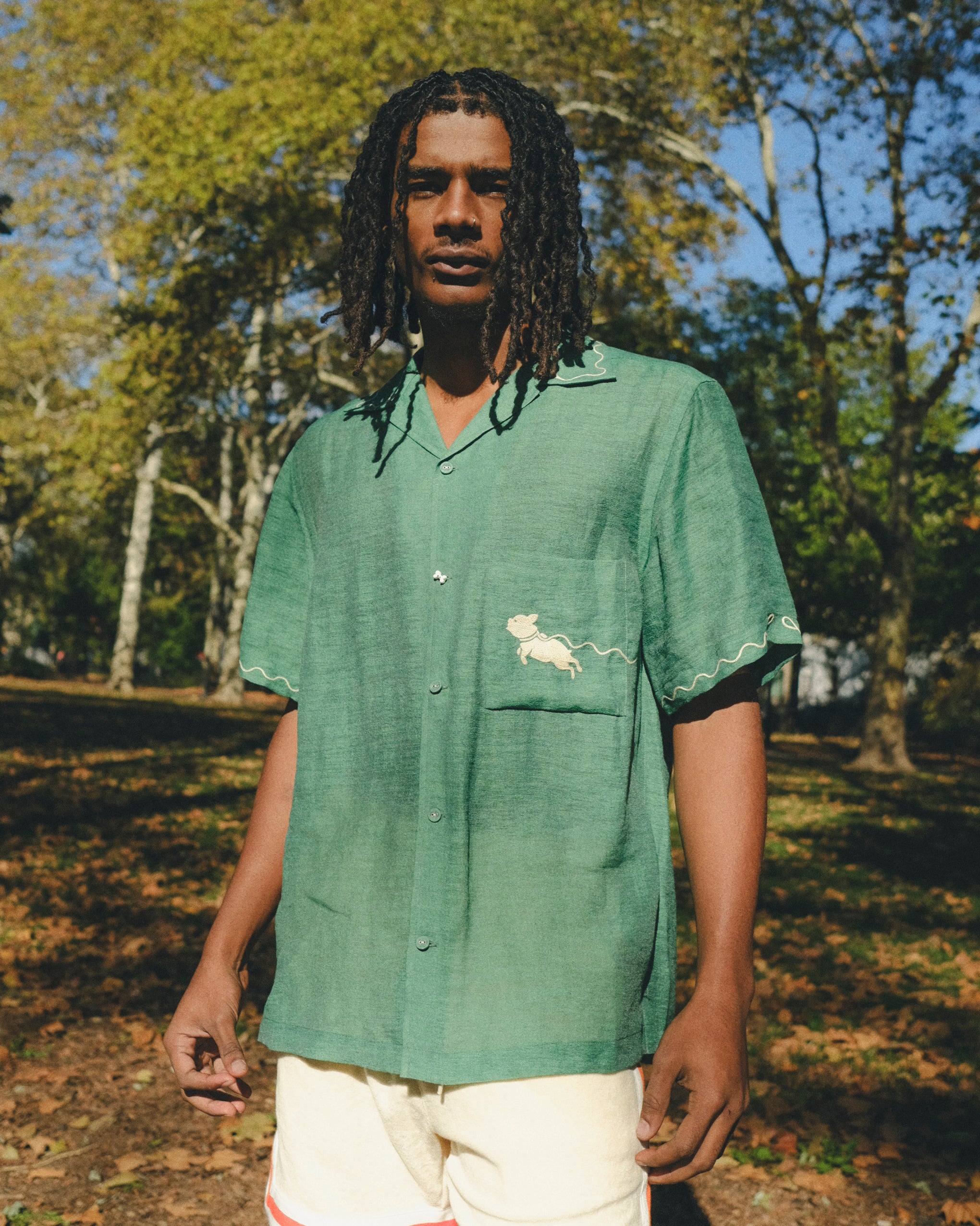 Man wearing green cabana shirt with contrast wavy piping, chest pocket with embroidered dog graphic, and a dog bone button at the center front.