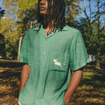 Man wearing green cabana shirt with contrast wavy piping, chest pocket with embroidered dog graphic, and dog bone button at the center front.