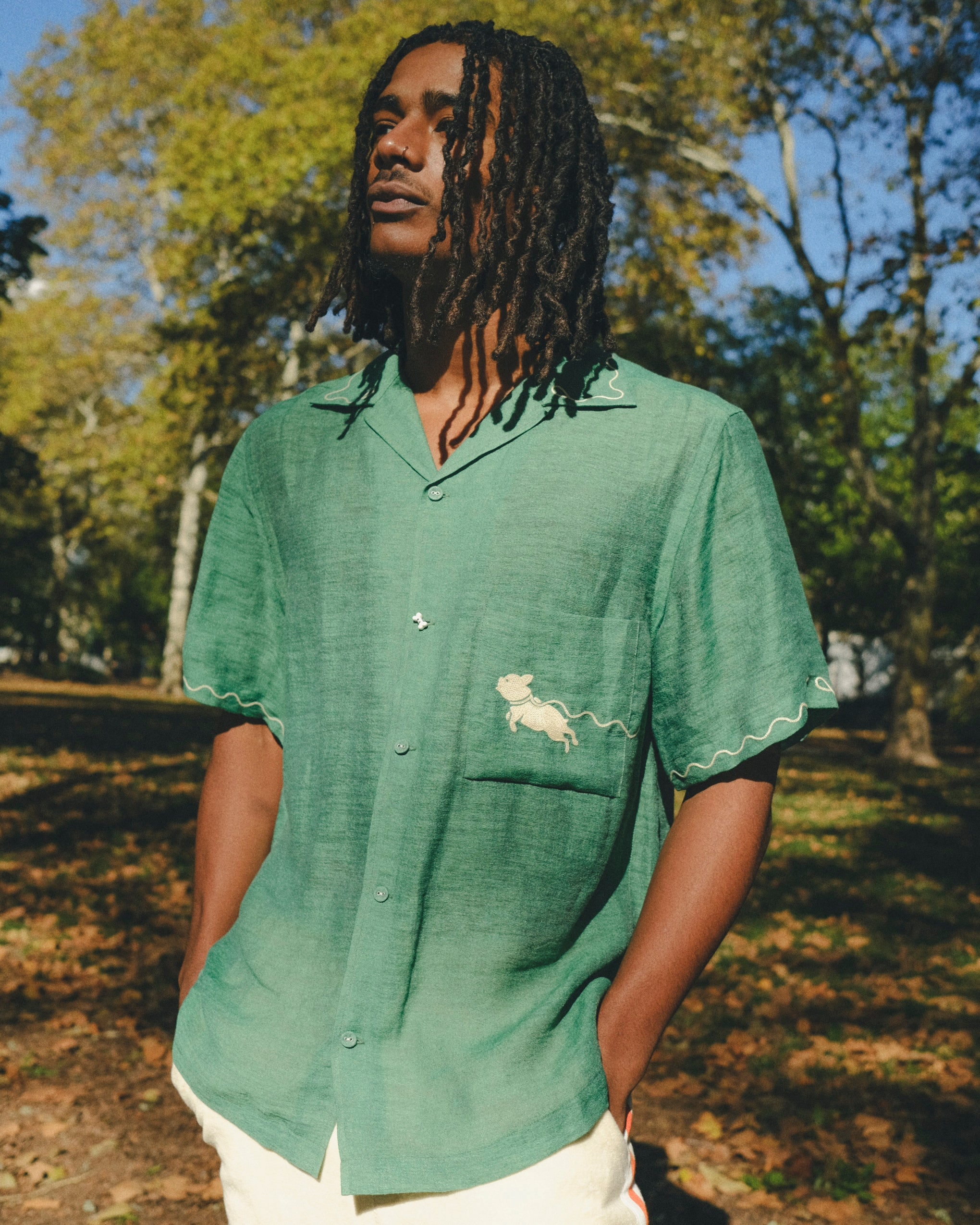Man wearing green cabana shirt with contrast wavy piping, chest pocket with embroidered dog graphic, and dog bone button at the center front.