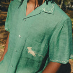 Close-up of man wearing green cabana shirt with contrast wavy piping along the collar and sleeves, chest pocket with embroidered dog graphic, and dog bone button at the center front.