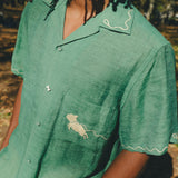 Close-up of man wearing green cabana shirt with contrast wavy piping along the collar and sleeves, chest pocket with embroidered dog graphic, and dog bone button at the center front.