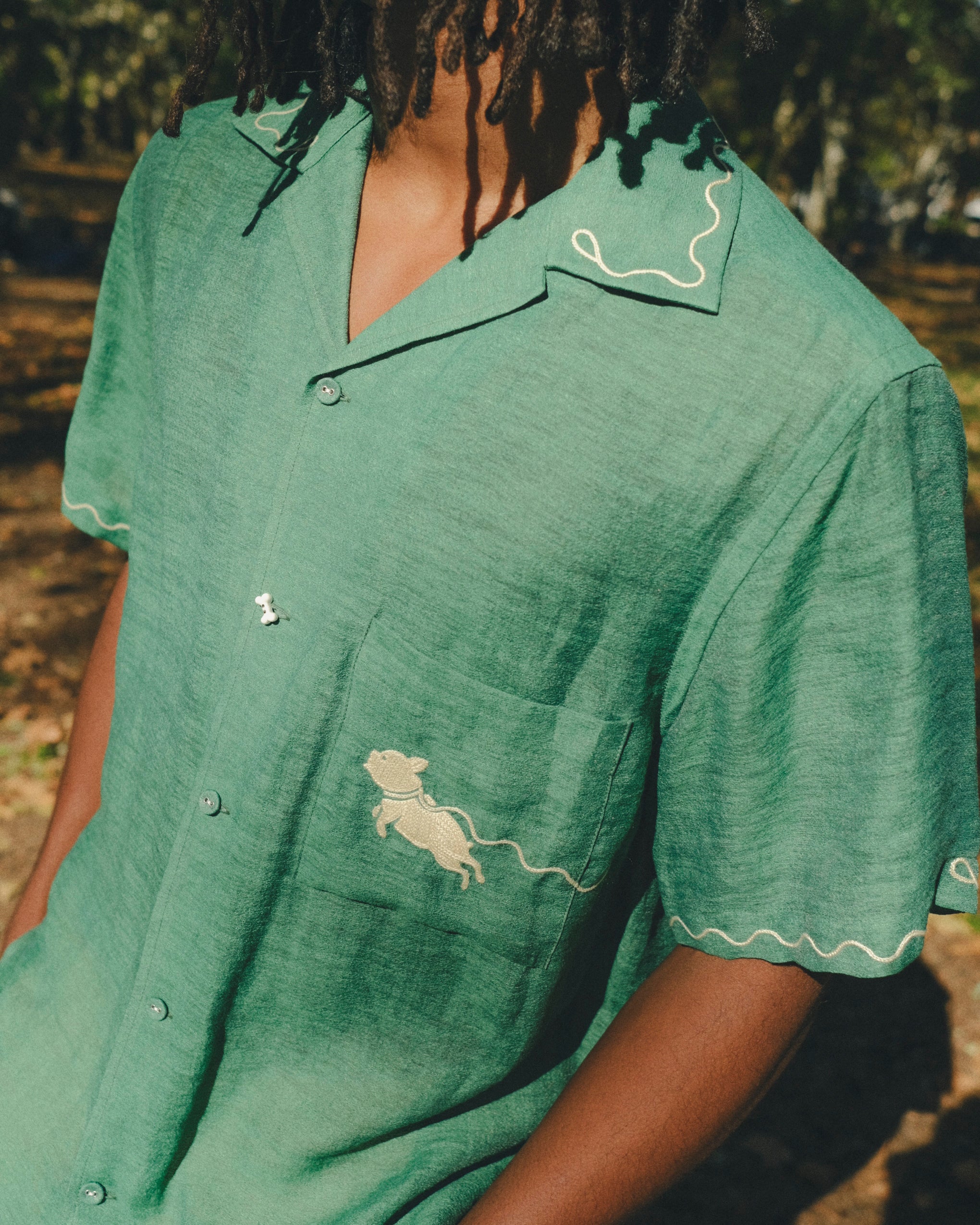 Close-up of man wearing green cabana shirt with contrast wavy piping along the collar and sleeves, chest pocket with embroidered dog graphic, and dog bone button at the center front.