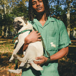 Man wearing green cabana shirt with contrast wavy piping, chest pocket with embroidered dog graphic, and dog bone button at the center front while holding a small dog.