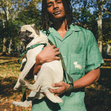Man wearing green cabana shirt with contrast wavy piping, chest pocket with embroidered dog graphic, and dog bone button at the center front while holding a small dog.