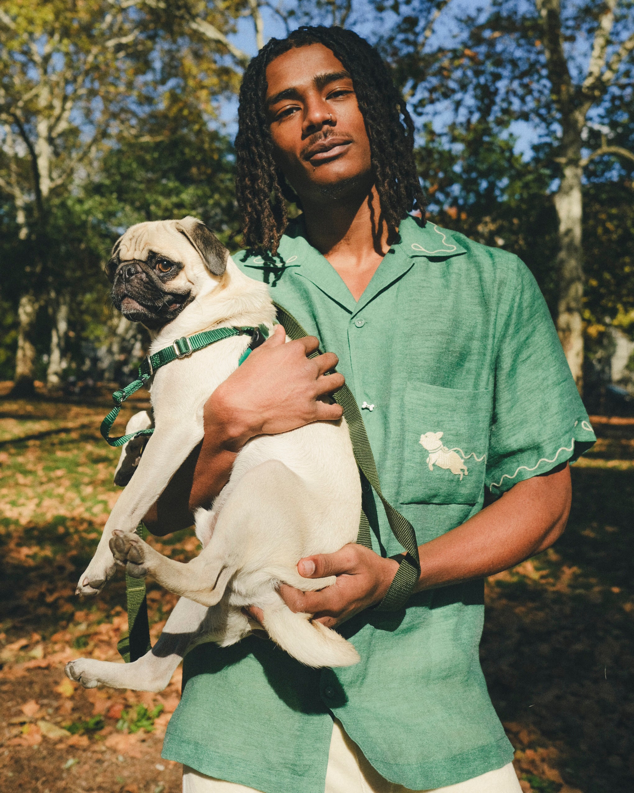 Man wearing green cabana shirt with contrast wavy piping, chest pocket with embroidered dog graphic, and dog bone button at the center front while holding a small dog.