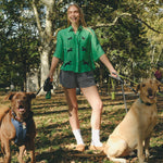 Woman wearing a green cropped cabana shirt with black dog silhouettes and white leash embroidery, standing with two dogs on leashes.