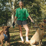 Woman wearing a green cropped cabana shirt with black dog silhouettes and white leash embroidery, standing with two dogs on leashes.