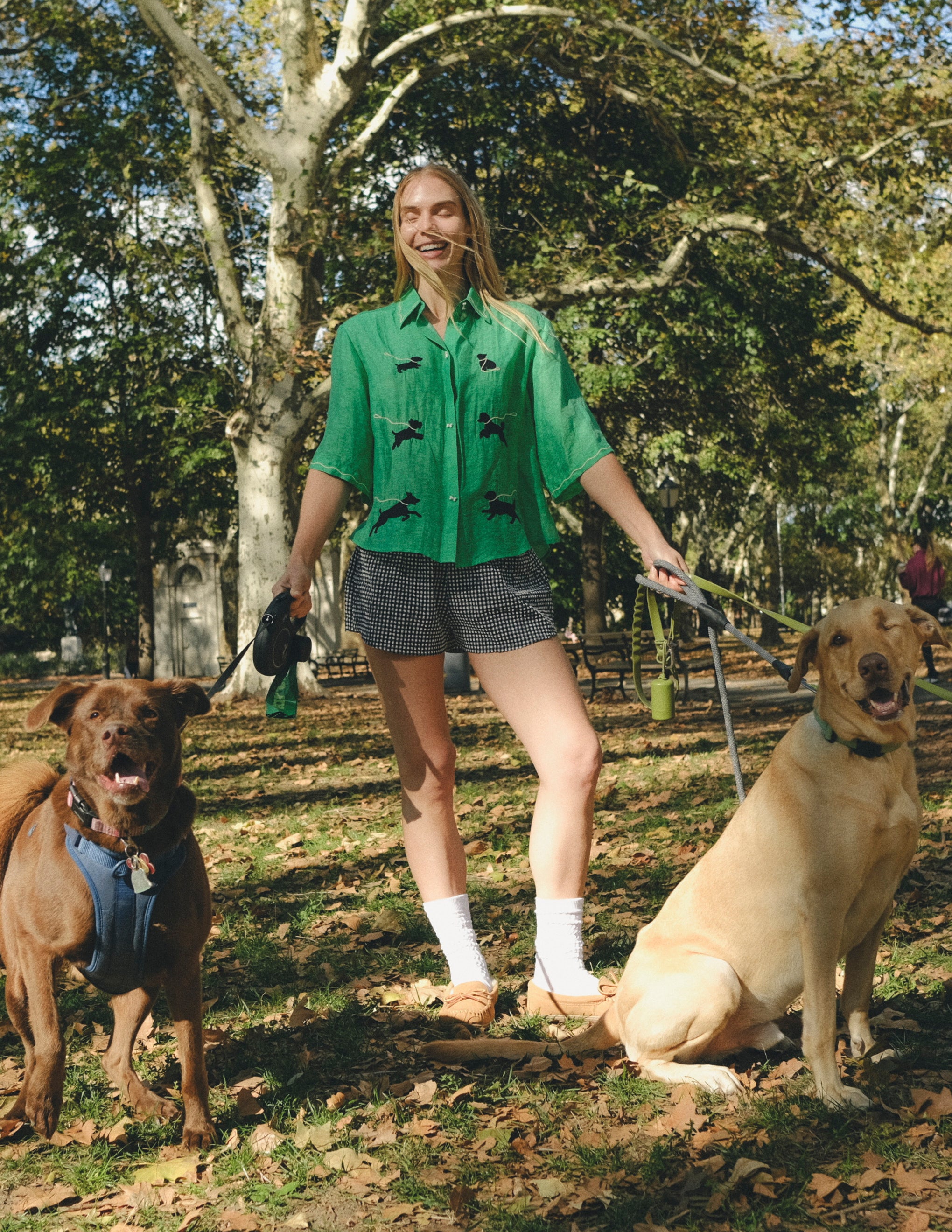 Woman wearing a green cropped cabana shirt with black dog silhouettes and white leash embroidery, standing with two dogs on leashes.