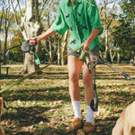 Woman wearing a green cropped cabana shirt with black dog silhouettes and white leash embroidery while holding two dog leashes.