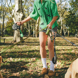 Woman wearing a green cropped cabana shirt with black dog silhouettes and white leash embroidery while holding two dog leashes.