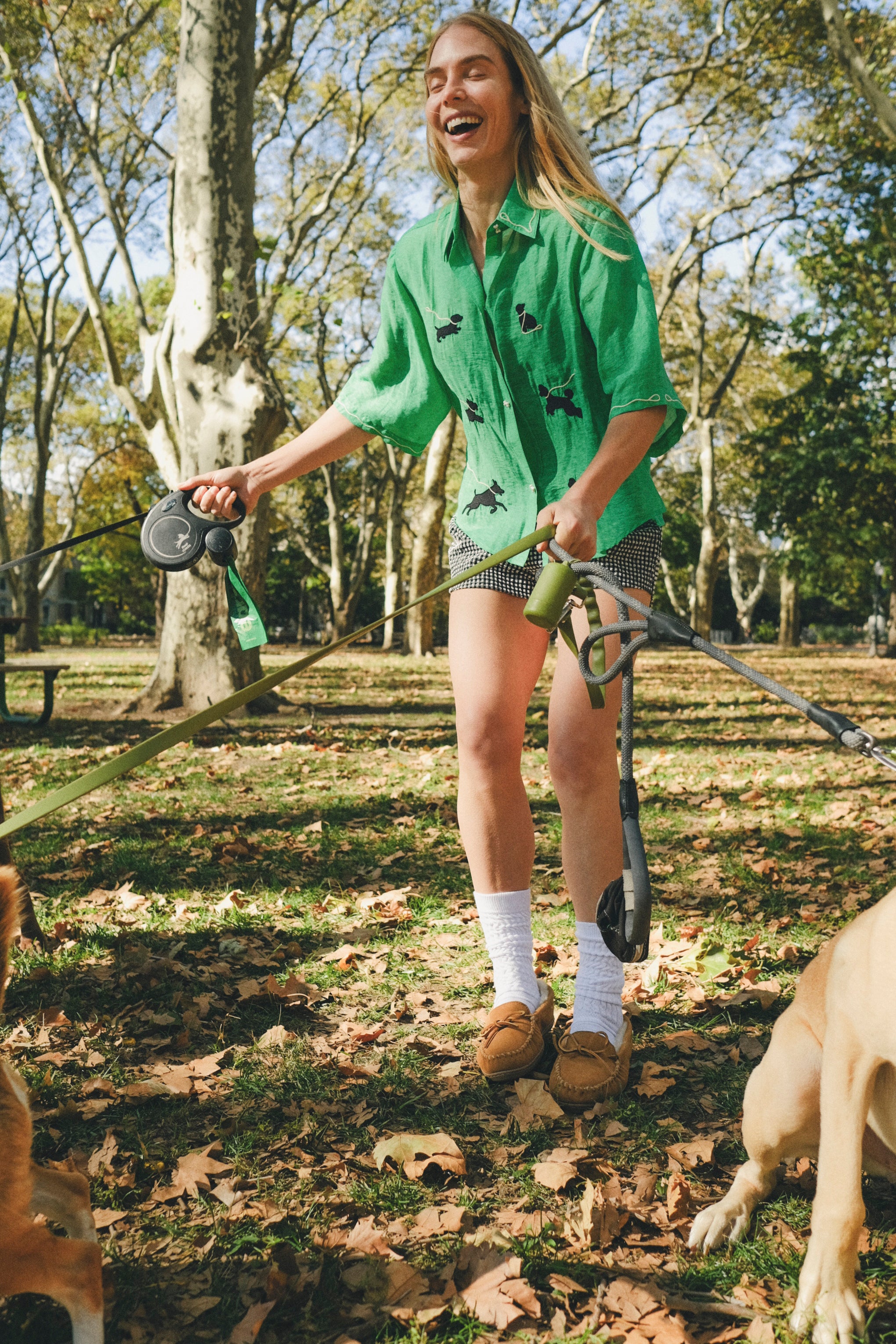 Woman wearing a green cropped cabana shirt with black dog silhouettes and white leash embroidery while holding two dog leashes.