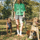 Woman wearing a green cropped cabana shirt with black dog silhouettes and white leash embroidery, standing with three dogs on leashes.