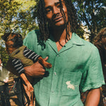 Man wearing green cabana shirt with contrast wavy piping, chest pocket with embroidered dog graphic, and dog bone button at the center front while holding a small dog.