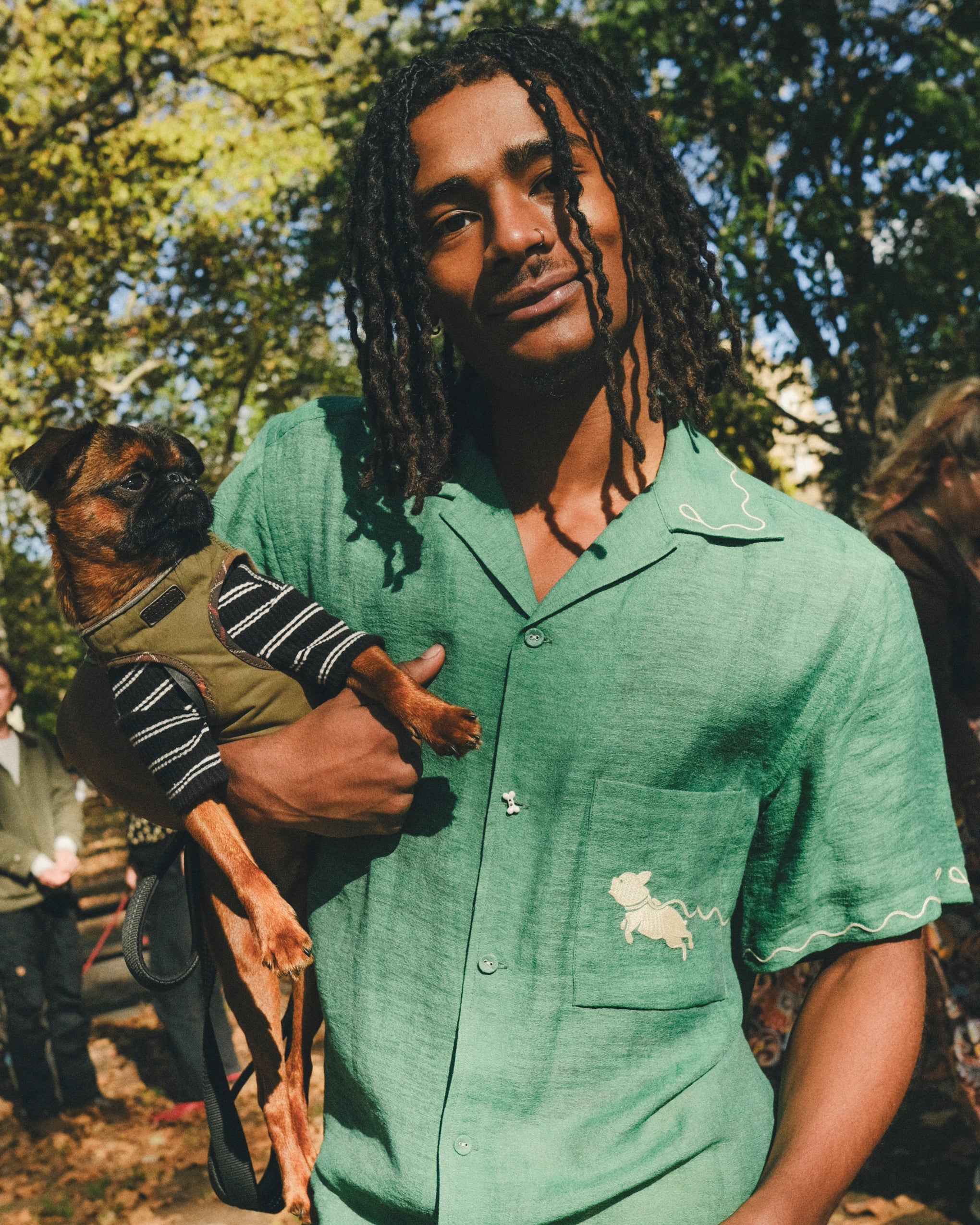 Man wearing green cabana shirt with contrast wavy piping, chest pocket with embroidered dog graphic, and dog bone button at the center front while holding a small dog.