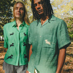 Two models wearing green cabana shirts with embroidered dog graphics, contrast wavy piping, and dog bone button detail at the center front.