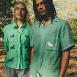Two models wearing green cabana shirts with embroidered dog graphics, contrast wavy piping, and dog bone button detail at the center front.