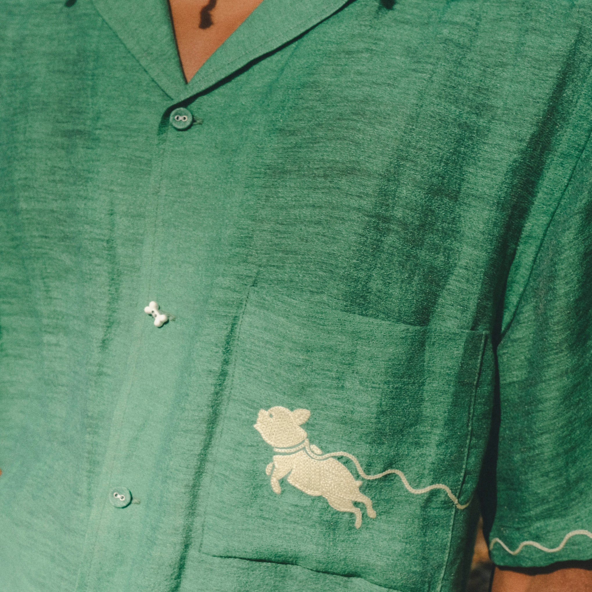 Close-up of green cabana shirt showing chest pocket with embroidered dog graphic, contrast wavy piping on the sleeve, and dog bone button at the center front.