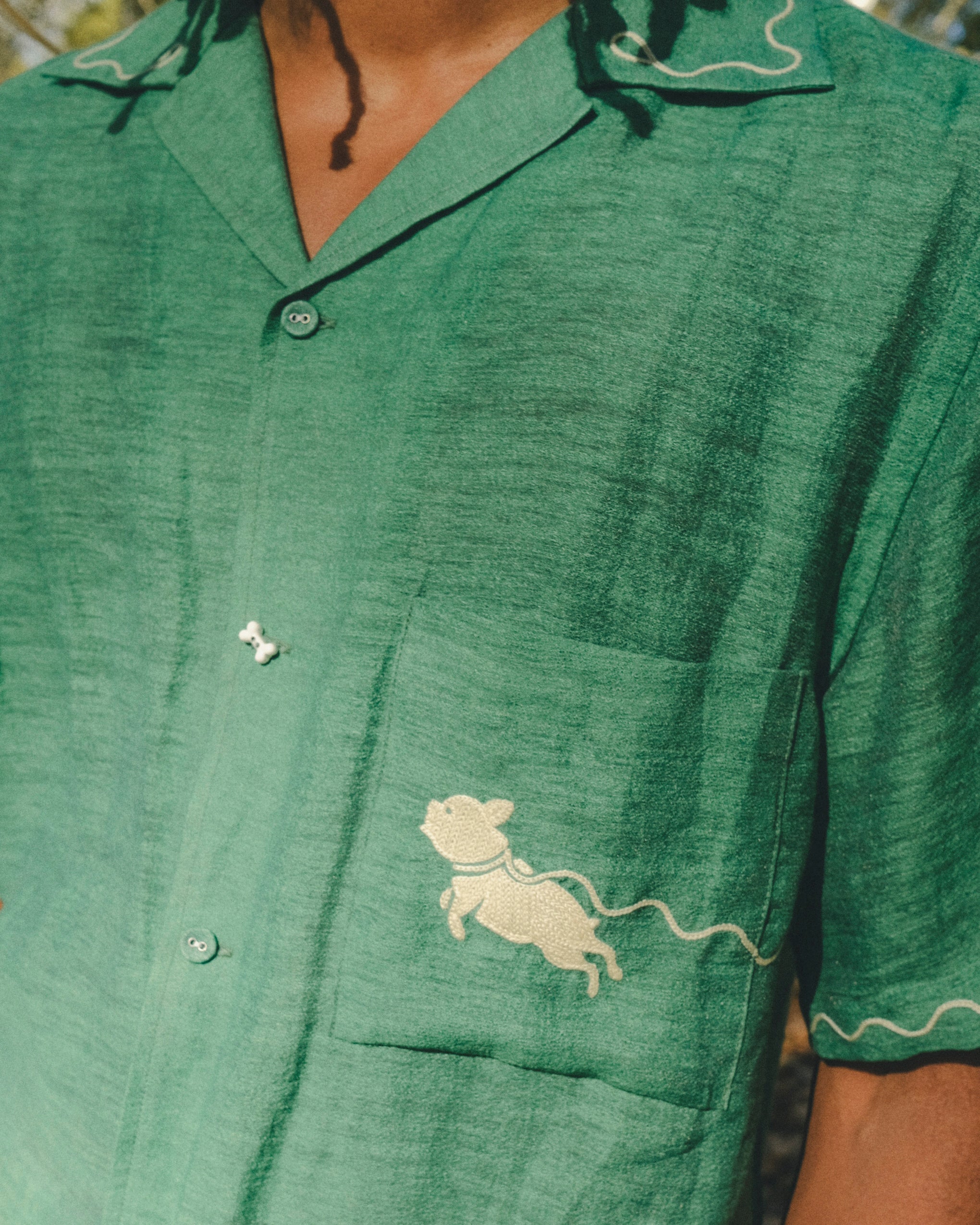 Close-up of green cabana shirt showing chest pocket with embroidered dog graphic, contrast wavy piping on the sleeve, and dog bone button at the center front.