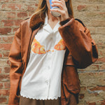 Woman in a white seersucker shirt with croissant embroidery and brown shorts, wearing a brown leather jacket. 