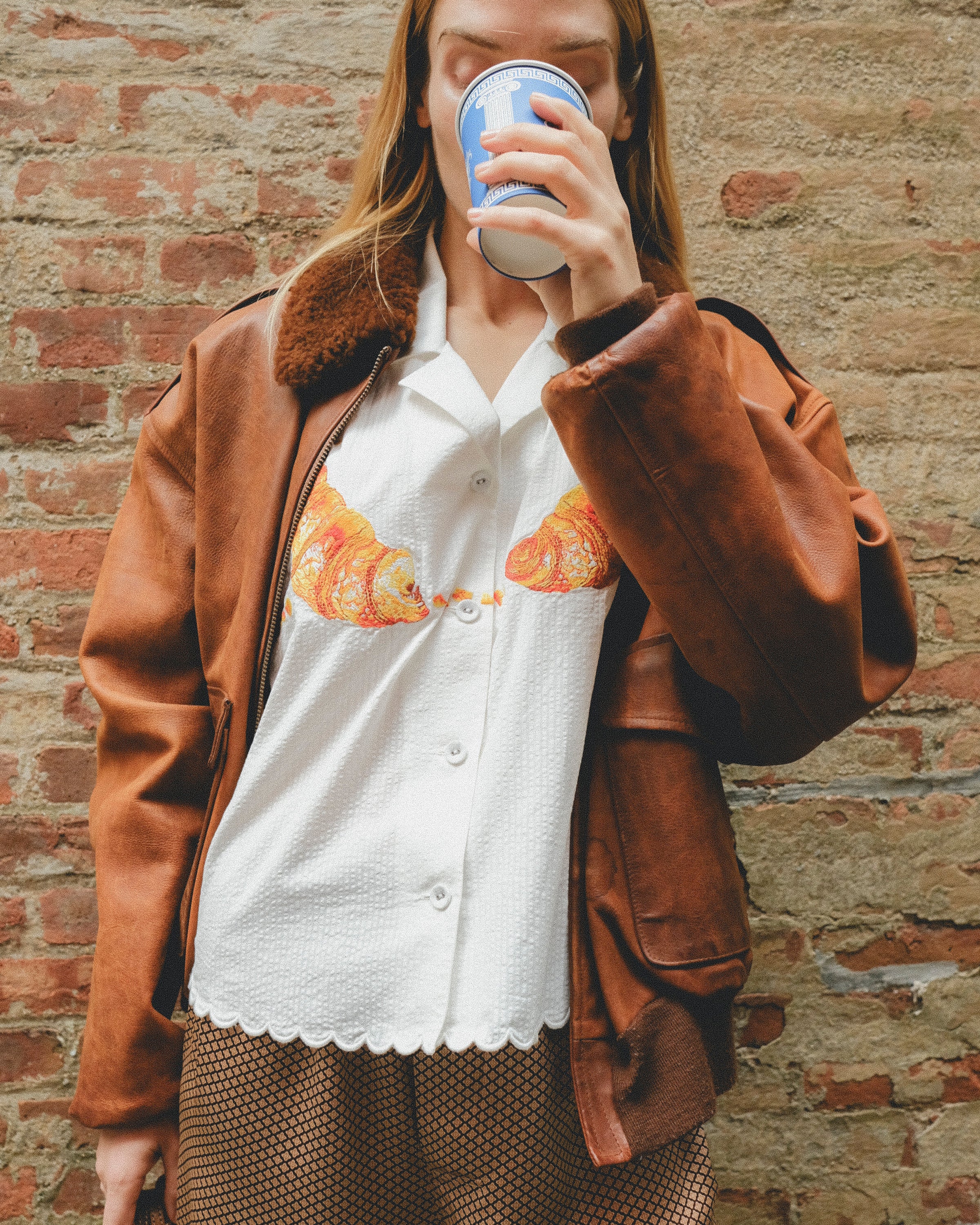 Woman in a white seersucker shirt with croissant embroidery and brown shorts, wearing a brown leather jacket. 